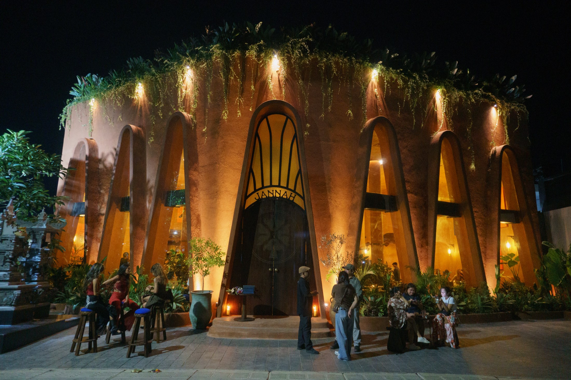 Jannah Restaurant in Seseh, Bali: a terracotta building with tall lit arches and a hanging garden roof at night